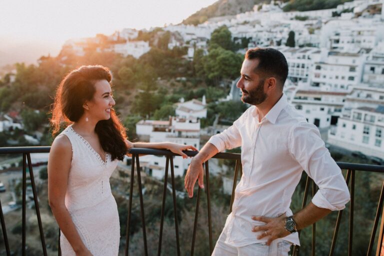 Casal vestido de branco se olhando. Ao fundo podemos observar uma paisagem com varias casinhas brancas que se misturam com a vegetação da Serra de Mijas