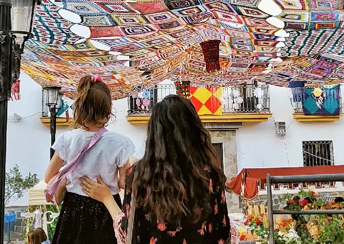 Mãe e filha em um mercadinho medieval com a rua coberta por um grande toldo de crochê durante o Festival Luna Mora de Guaro.